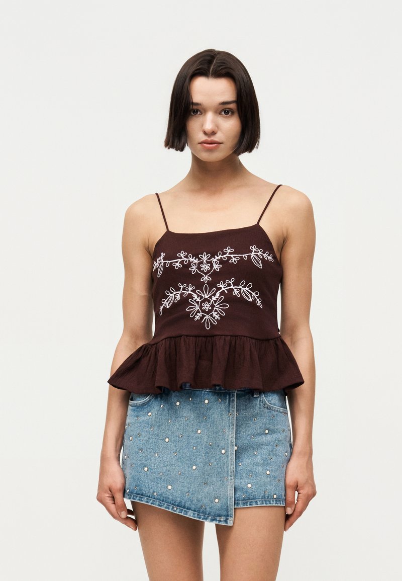 Femme aux cheveux courts et foncés portant un top spaghetti marron foncé avec une broderie florale blanche et une jupe en jean asymétrique cloutée.