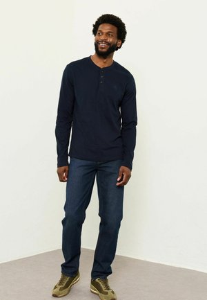 Lächelnder Mann mit Afro-Haaren, der ein marineblaues Langarm-Henley-Shirt, blaue Jeans und olivgrüne Sneaker trägt und vor einer schlichten Wand steht.