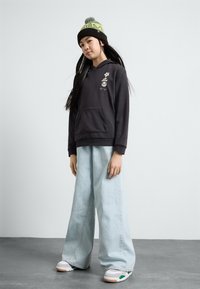 Hoodie noir avec broderie florale, poche avant, associé à un jean large bleu clair et des baskets blanches sportives avec des accents verts.