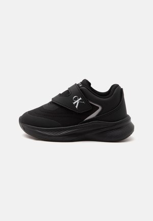 Baskets noires Calvin Klein avec fermeture à scratch, logo cK blanc sur la bride, et semelle épaisse texturée sur fond blanc.