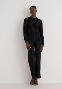 Schwarze, durchsichtige Bluse mit langen Ärmeln und Knopfleiste, kombiniert mit hochgeschnittenen schwarzen Hosen und schwarzen Sandalen mit Riemchen. Schlichtes, tailliertes Design.