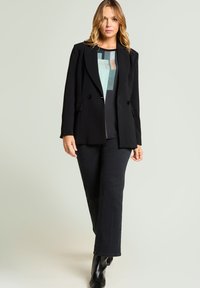 Schwarzer zweireihiger Blazer mit Reverskragen, getragen über einem gemusterten Oberteil, kombiniert mit schwarzen Schlaghosen und eleganten schwarzen Stiefeln.