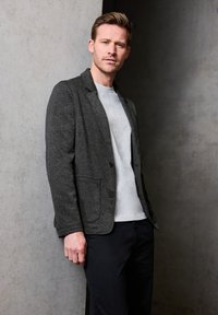 Szary wełniany żakiet z dwoma przednimi kieszeniami i zapięciem na guziki, noszony na jasnoszarym t-shircie, w połączeniu z ciemnymi spodniami na tle betonowym.