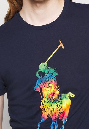 T-shirt en coton bleu marine avec un grand graphique abstrait de joueur de polo coloré, mêlant des couleurs arc-en-ciel et des accents noirs.