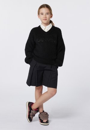 Pull en tricot noir avec un col en V et un logo, associé à un short plissé noir. Porté avec des baskets uniques avec des lacets et des accents à motifs.