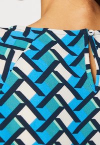 Blå mønstrede kjolestof har et geometrisk design med sammenflettede former i nuancer af blå, grøn og hvid. Detalje med knaplukning bagpå er synlig.