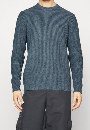 Homme portant un pull en maille texturée bleu à manches longues et un pantalon cargo noir, se tenant avec les bras détendus le long du corps.