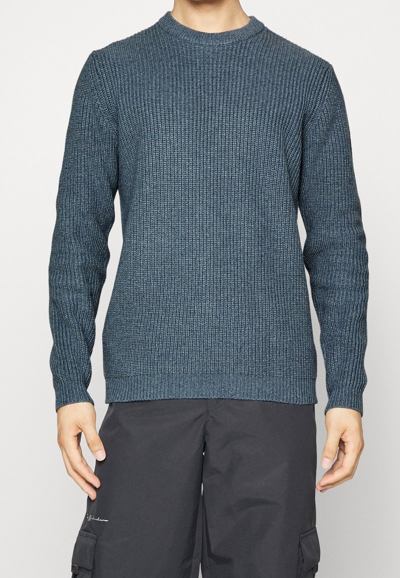 Homme portant un pull en maille texturée bleu à manches longues et un pantalon cargo noir, se tenant avec les bras détendus le long du corps.