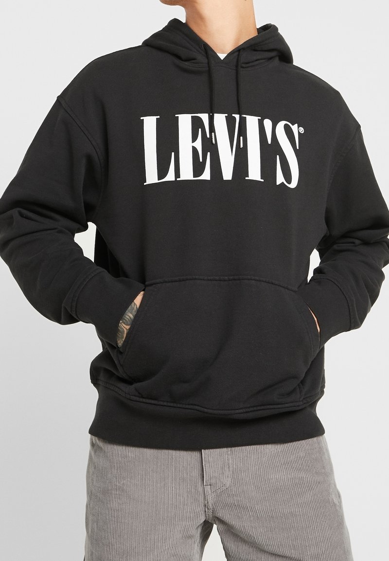 Personne portant un sweat à capuche noir Levi's avec un logo texte blanc, les mains dans la poche avant, assorti à un pantalon en velours côtelé gris clair.