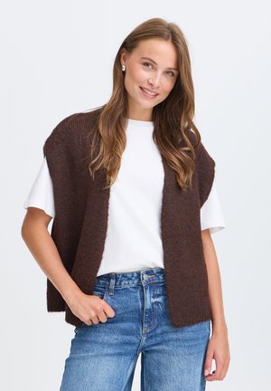 Jonge vrouw met lang bruin haar draagt een wit t-shirt, een bruine mouwloze gebreide vest en een blauwe jeans, glimlachend tegen een effen achtergrond.