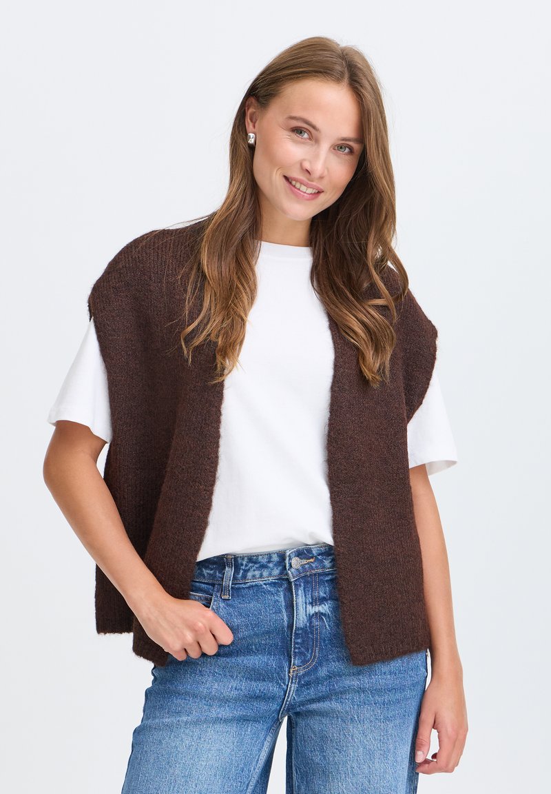 Jeune femme aux longs cheveux bruns portant un t-shirt blanc, un gilet en maille marron sans manches et un jean bleu, souriante devant un fond uni.