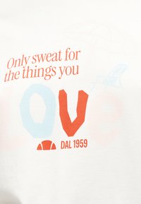 Κείμενο σε κόκκινο και μπλε χρώμα σε λευκό ύφασμα που γράφει «Μόνο ιδρώστε για τα πράγματα που αγαπάτε» με ένα εικονίδιο μπάσκετ και το «DAL 1959» από κάτω.