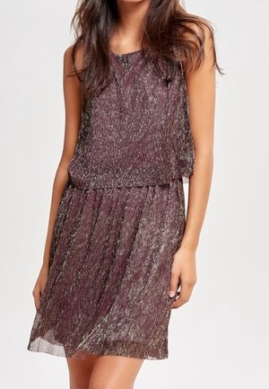 Kobieta w bezrękawną, fioletowo-srebrną, metaliczną sukienkę o teksturze, sięgającą do kolan, z luźną spódnicą i dopasowaną talią.