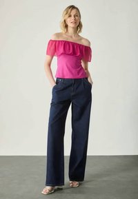 Ροζ τοπ με off-the-shoulder σχέδιο και λεπτομέρειες ρίγες, συνδυασμένο με φαρδιά σκούρα μπλε τζιν. Το μοντέλο στέκεται με τα χέρια στις τσέπες σε μια υφασμάτινη επιφάνεια.