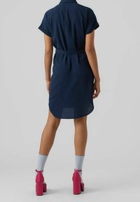 Vestito camicia blu navy con maniche corte arrotolate, vita accentuata e orlo curvo. Abbinato a scarpe con tacco a blocco rosa e calze azzurre chiare.