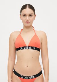 Pomarańczowy stanik bikini z prążkowanej tkaniny z czarną bandą z logo Calvin Klein, z trójkątnym fasonem i ramiączkami na szyi; w komplecie również dopasowane majtki bikini.