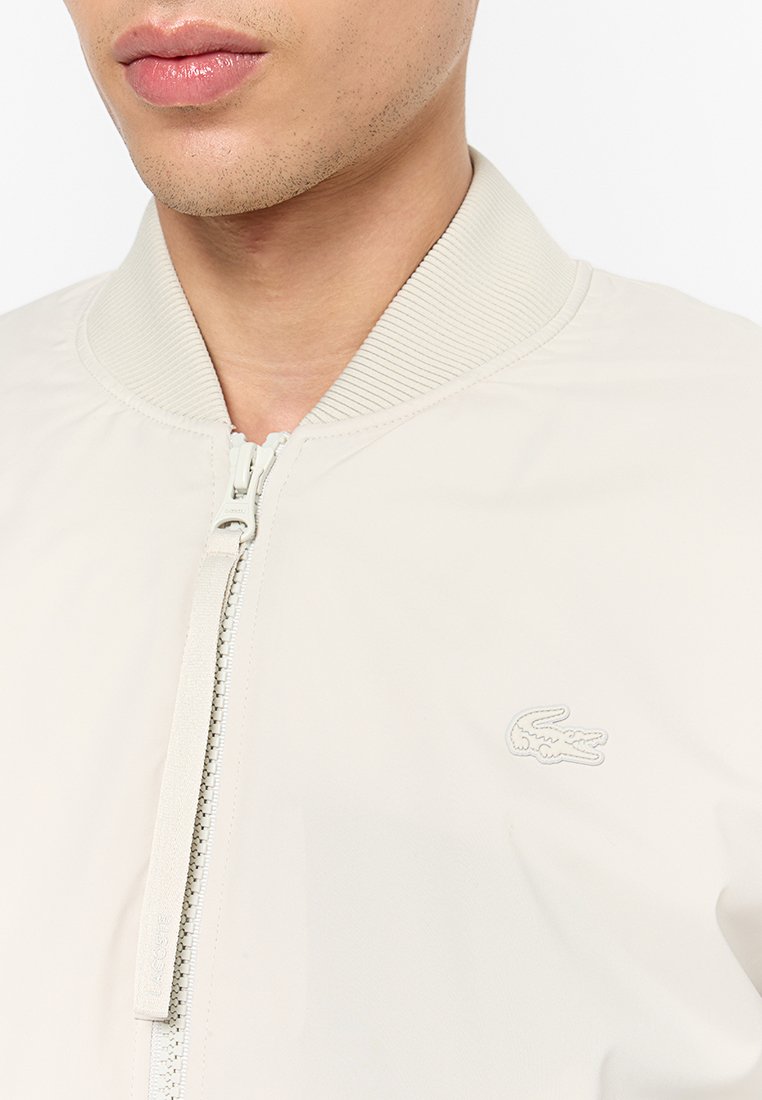 Primo piano di una persona che indossa una giacca beige chiara con zip, colletto a coste e logo di coccodrillo sul petto.