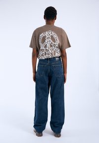 T-shirt grafica marrone con un grande segno di pace floreale bianco sulla schiena, abbinato a jeans larghi blu scuro.