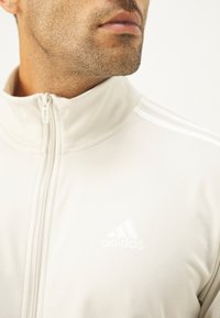 Biely zipsový sveter s vysokým golierom, s tromi bielymi pruhmi na rukávoch a vyšitým logom Adidas na hrudi.