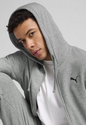 Lys grå zip-up hættetrøje lavet af blødt stof, med en kengurulomme og et sort Puma logo på venstre bryst. Hvid t-shirt under.