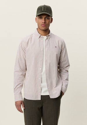 Homme portant une chemise à rayures boutonnée par-dessus un t-shirt blanc, un pantalon foncé et une casquette vert foncé avec un logo sur le devant, debout devant un fond uni.
