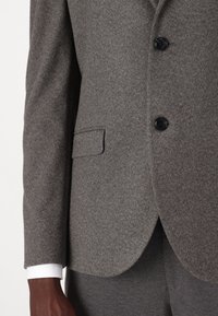 Mörkhyad person klädd i en grå texturerad kavaj med svarta knappar och matchande byxor, vit skjortärm synlig.