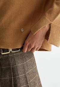 Maglione lavorato a maglia color cammello con orlo a coste e spacco sulle maniche, dotato di un piccolo dettaglio logo in argento. Abbinato a una gonna a quadri marrone e una cintura.