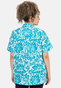 Camicia a maniche corte con un motivo floreale blu e verde su sfondo bianco, con colletto button-down e dettagli sul giromanica.