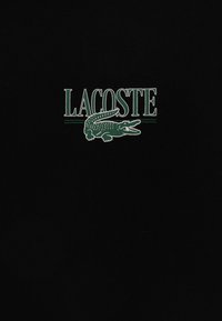 Svart bomull T-skjorte med ordet "LACOSTE" og en grønn krokodillelogo, sentrert på brystet med vertikale aksenter.