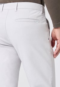 Pantaloni grigi chiari con una texture liscia, dotati di una tasca laterale e un'etichetta con logo. Progettati con una vestibilità slim e un sottile dettaglio cucito.