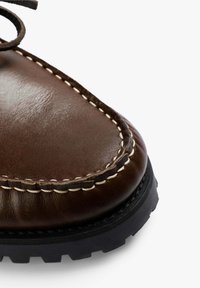 Zapato de cuero marrón con un acabado pulido, que presenta costuras blancas a lo largo del borde y una suela de goma negra texturizada.
