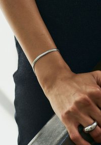 Bracciale d'argento dal design liscio e flessibile indossato al polso. Il bracciale presenta una forma sottile e cilindrica con una texture delicata.