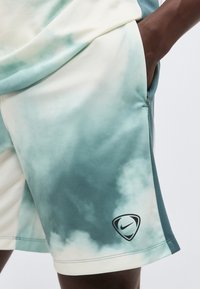 Κοντινή λήψη ανοιχτό μπλε και λευκών βερμούδων tie-dye με μαύρο λογότυπο Nike στο κάτω πόδι, φορεμένα από άτομο με το χέρι στην τσέπη.