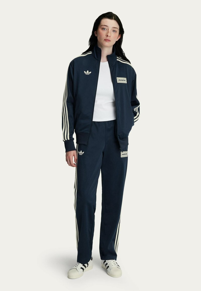 Navy tracksuit with white stripes, fitted jacket and pants, featuring two logos. Worn with a white shirt and classic sneakers.