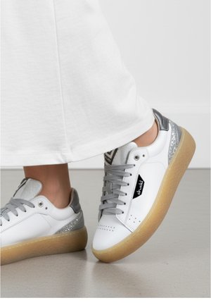 Zapatilla blanca de cuero con acentos grises, parte trasera texturizada y cordones metálicos. Cuenta con una suela de goma y pequeñas perforaciones en la puntera.