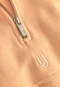 Perzikkleurige ritsjack met een zilveren rits en geborduurd logo. Glad textuur met stofdetails nabij de rits.