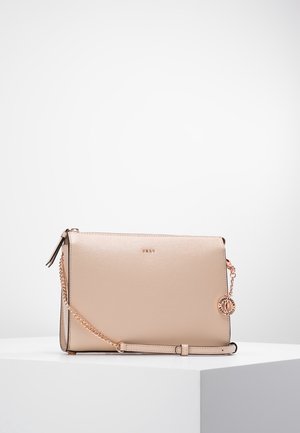 Bolso bandolera de cuero beige con cadena de metal dorada y colgante circular con logo metálico, colocado sobre una superficie blanca contra un fondo liso.