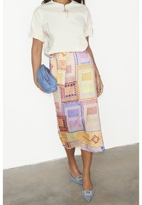 Weißes, kurzärmliges Oberteil mit Applikationsdetails, kombiniert mit einem bunten, gemusterten Midirock mit geometrischen Formen. Blaue Clutch und High Heels.