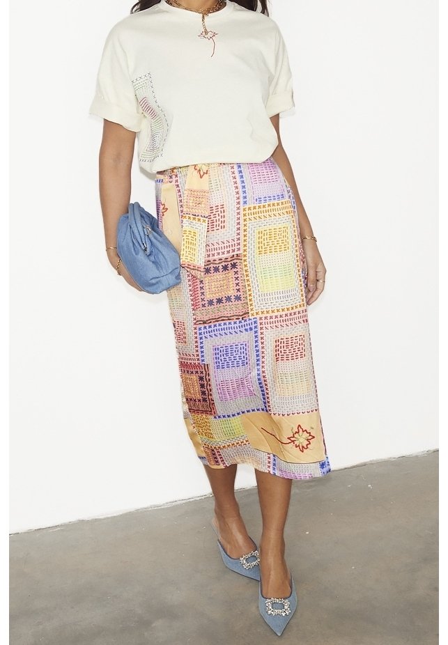 Weißes, kurzärmliges Oberteil mit Applikationsdetails, kombiniert mit einem bunten, gemusterten Midirock mit geometrischen Formen. Blaue Clutch und High Heels.