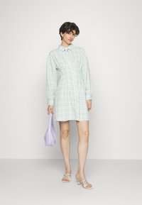 Vestido camisa em padrão xadrez verde claro, com mangas longas, Botões na parte da frente e colarinho. Combinado com uma mala lavanda e sapatos de salto bege.