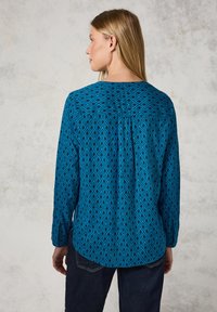Camicia azzurra a maniche lunghe con motivo a rombi, dotata di spalla posteriore e dettagli con pieghe. Abbinata a jeans scuri.