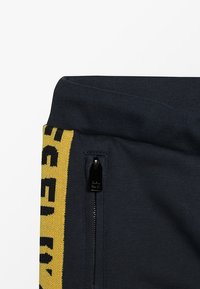 Mörkgrå träningsbyxor med en gul sidremsa med svart text, dragkedjeficka och ribbad midjeväv.