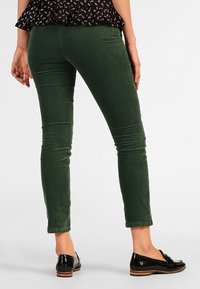Vrouw in donkere groene slim-fit broek en zwarte gepatineerde loafers, staand met handen ontspannen langs haar zij op een effen achtergrond.