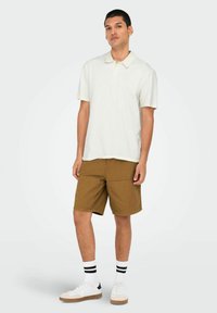 Cremefarvet polo skjorte med korte ærmer, lyse shorts og hvide sneakers med brune detaljer. Model står imod en lys grå baggrund.