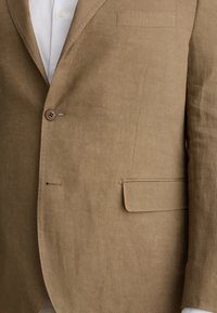 Blazer in lino beige con chiusura a un bottone, rever a punta, una tasca sul petto e una tasca laterale. Texture liscia, vestibilità sartoriale.