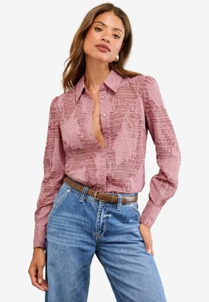 Rosa Bluse mit strukturierten Blattmustern, mit Knöpfen und langen Ärmeln. Kombiniert mit hochgeschnittenen blauen Jeans und einem braunen Gürtel.
