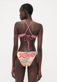Haut de bikini rose avec un design à dos entrelacé ; le bas présente un motif orange et gris avec le texte "Heart" et des textures contrastantes.