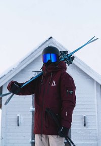 Bordo narciarska kurtka z czarnymi akcentami i logo, czarny kask, okulary z niebieskim odcieniem, trzymający narty i kijki narciarskie na tle zaśnieżonego krajobrazu.