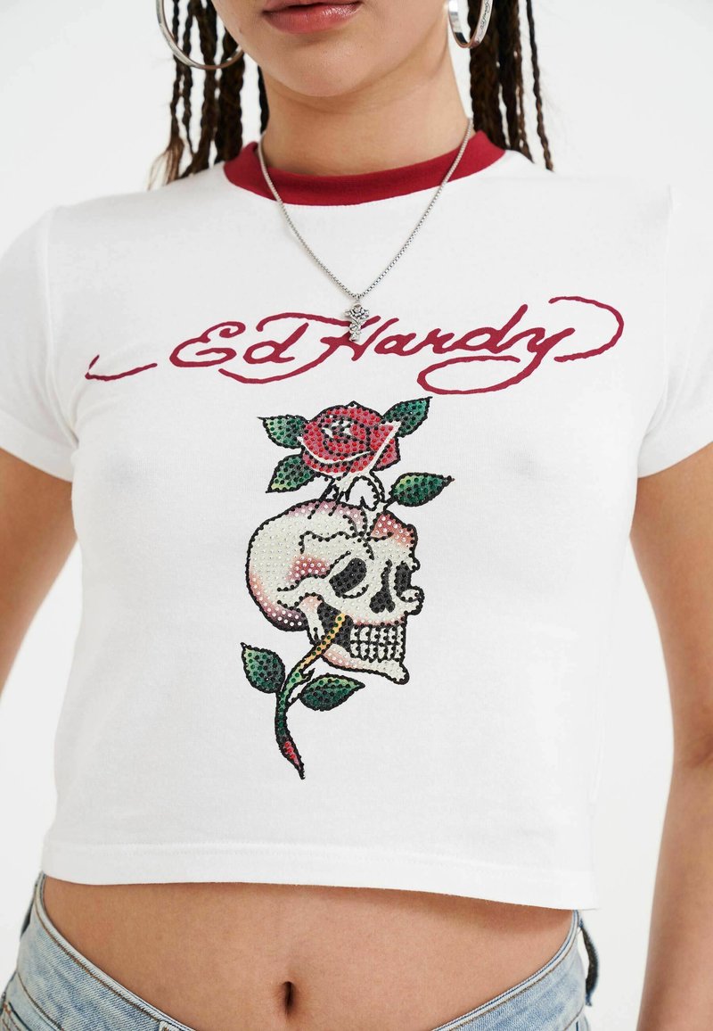 T-shirt bianca corta con colletto rosso e grafica di un teschio e una rosa in strass. Testo "Ed Hardy" in rosso sopra il design.