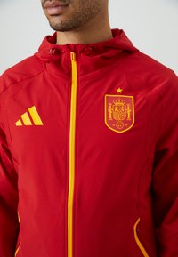 Giacca rossa con cappuccio e accenti gialli. Il fronte presenta una zip gialla e un logo con una corona. Realizzata in un materiale liscio e leggero.